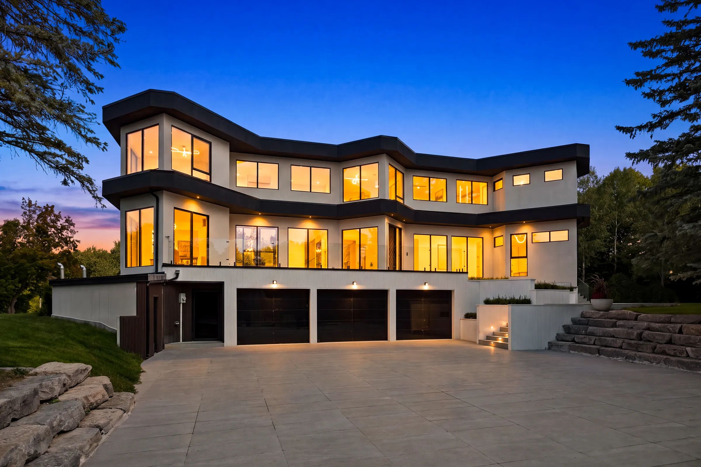 Stepped white facade at dusk, every window glowing warm and downlights washing the garage forecourt