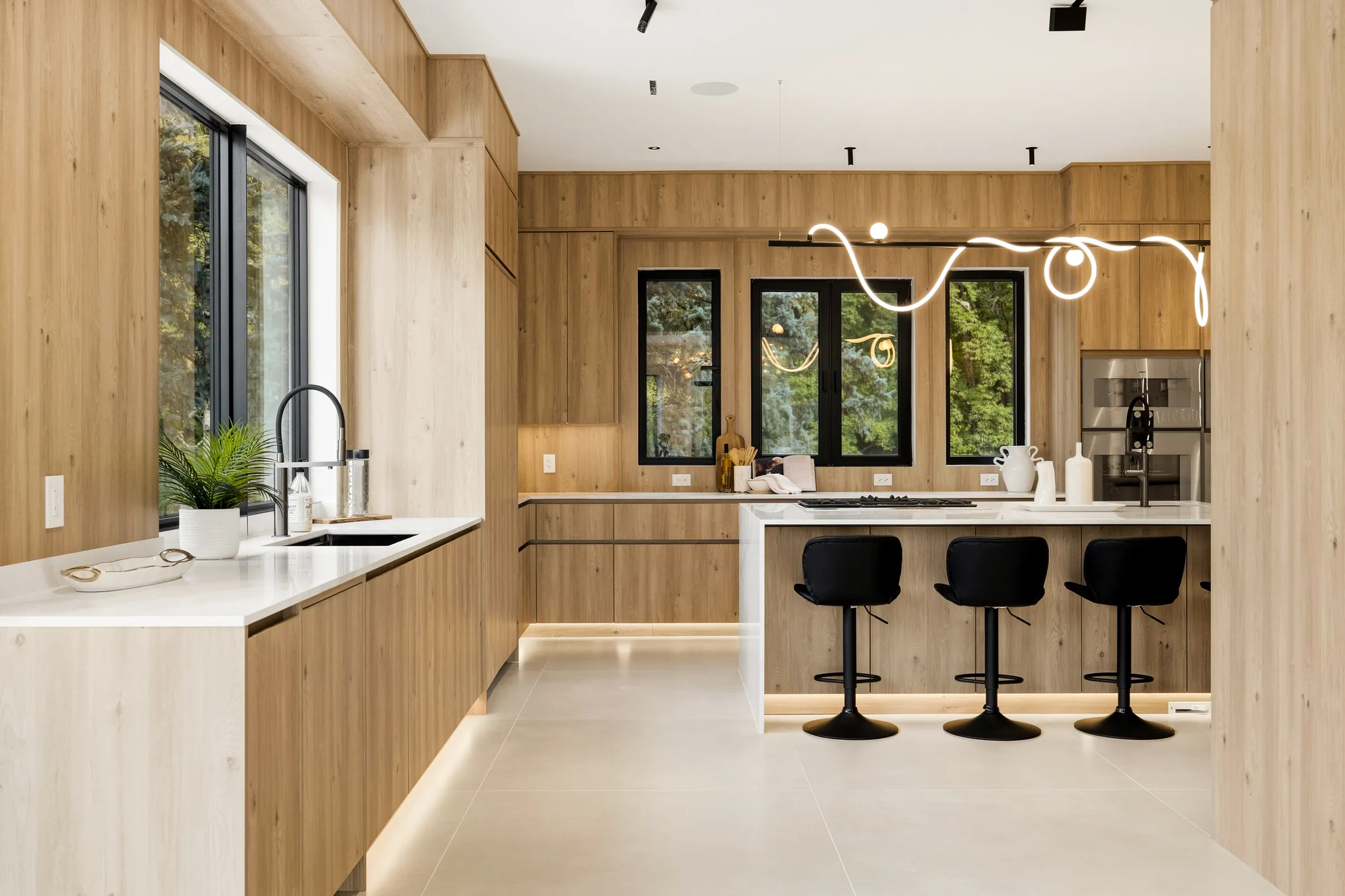 Kitchen with a looping light pendant over the island, toe kick lighting under the counters and black stools